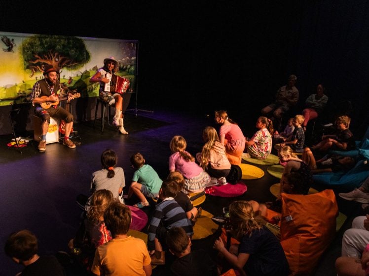 Image of Children sit on colourful cushions watching two musicians perform on stage in front of a backdrop with a tree illustration. One musician plays a ukulele, and the other plays an accordion. The audience looks engaged.