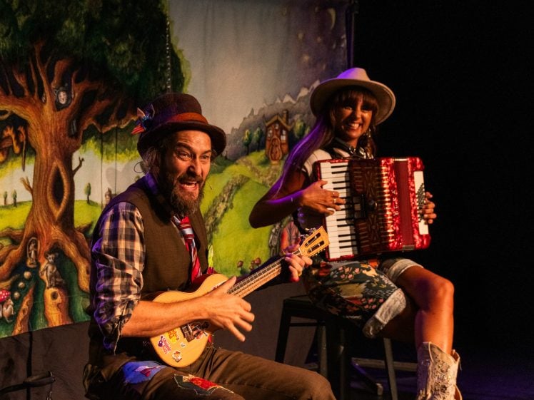 Image of Two performers in colourful costumes play instruments on stage; a man with a ukulele and a woman with an accordion, both smiling, with a whimsical, painted forest backdrop behind them.