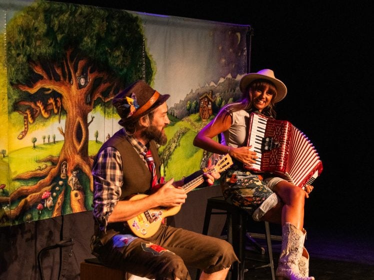 Image of A man playing a small guitar and a woman playing an accordion perform on stage, smiling, with a colourful, illustrated backdrop featuring a large tree and whimsical scenery behind them.