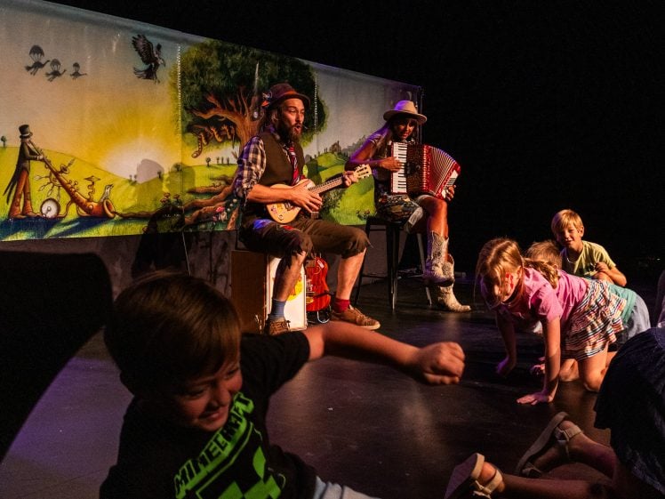 Image of Two musicians perform on stage, one playing a guitar and the other an accordion, in front of a colourful backdrop, while several children crawl and play energetically at their feet.