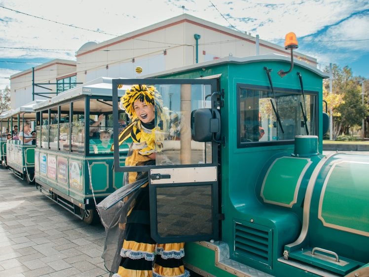 Image of A person dressed in a yellow and black bee costume cheerfully leans out of the door of a green trackless train parked on a paved area with buildings and trees in the background.