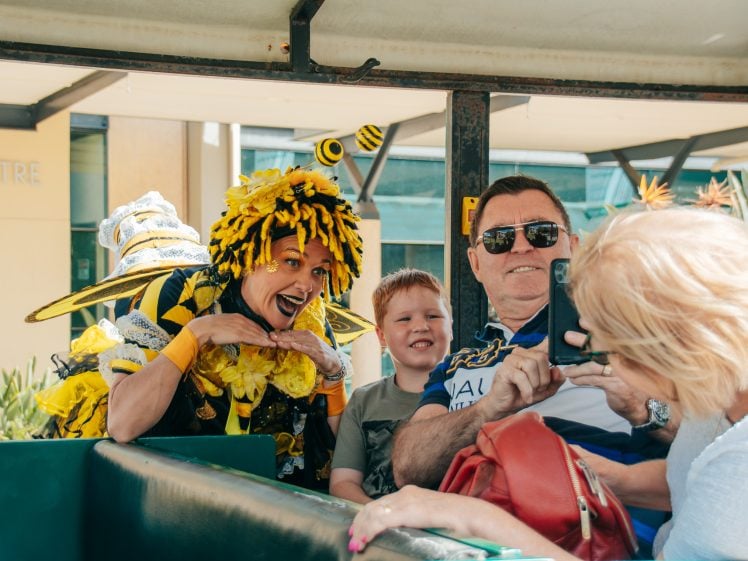 Image of A woman in a bright bee costume poses cheerfully next to a smiling boy and two adults, one of whom is taking a photo, as they sit together on an outdoor vehicle.