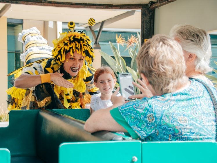 Image of A performer in a bee costume leans towards a smiling girl and two older women as one woman takes a photo on her mobile. They are all sitting in a green vehicle, enjoying the interaction outdoors.