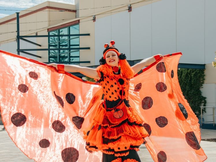 Image of A performer dressed in a vibrant ladybird costume with large red wings and black spots stands outside with arms spread wide, smiling brightly. The background features a modern building and some greenery.