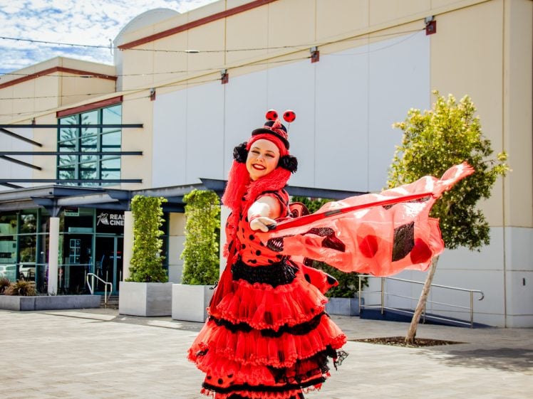 Image of A person dressed in a vibrant red and black ladybird-themed costume poses outdoors, smiling and holding a flowing scarf. The background features a modern building with greenery and large windows.