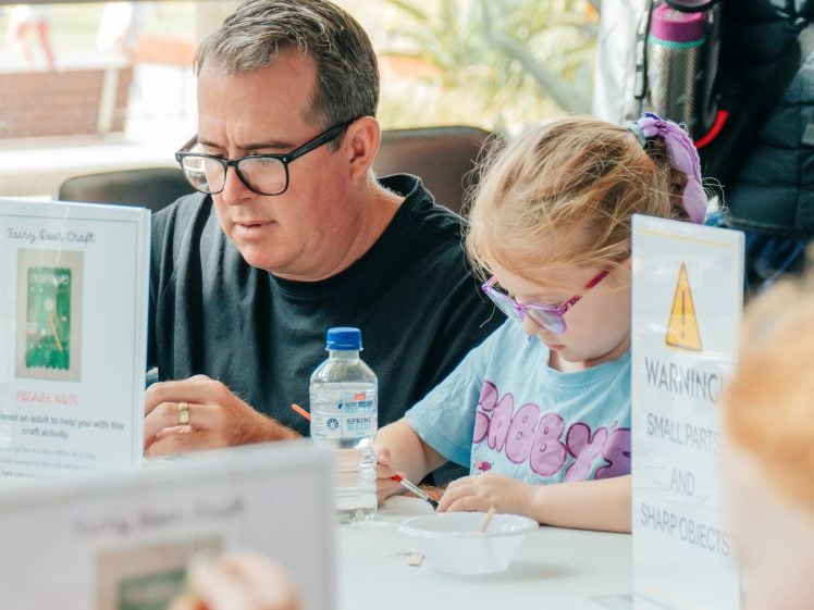 Image of A man and a young girl with glasses sit at a table doing a craft activity. Signs on the table warn about small parts and sharp objects. The girl is focused on her work, and a water bottle is placed nearby.