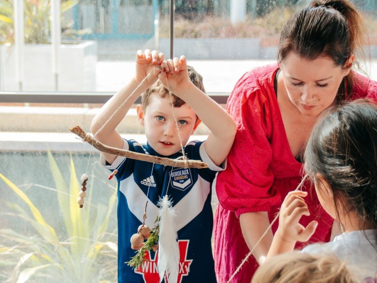 Image of A young boy in a blue sports shirt holds up a string with natural materials, while a woman in a pink top assists and other children work around them in a bright, sunlit room.