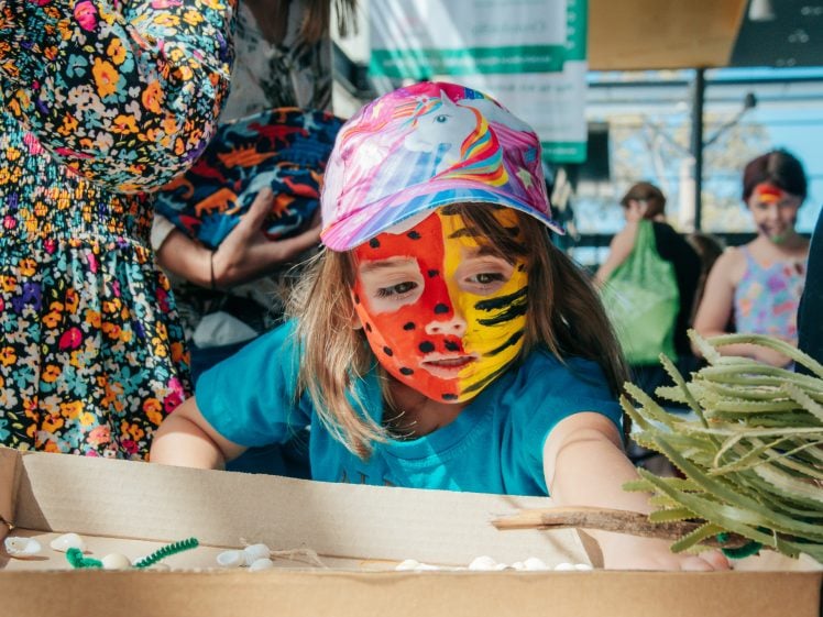 Image of A child with colourful animal face paint and a unicorn cap reaches into a box of craft materials, surrounded by people at a lively indoor event.