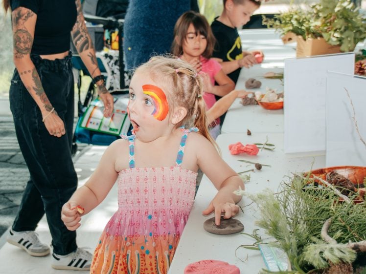 Image of A young girl with a rainbow painted on her face looks surprised whilst making crafts at a table with other children and an adult nearby. The table is covered with craft materials and plants.