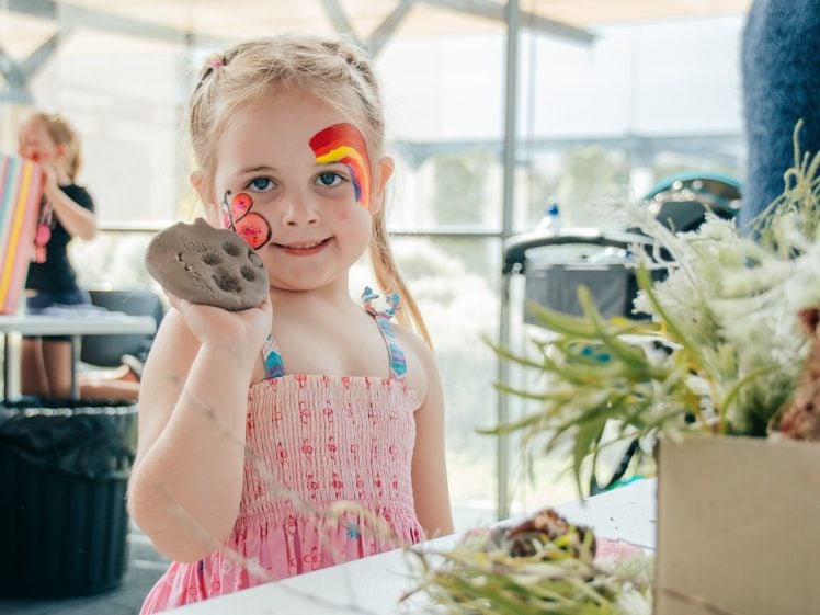 Image of A young girl with face paint and a rainbow on her cheek smiles whilst holding up a piece of clay at a table. She wears a pink dress, and there are plants and art supplies on the table in front of her.