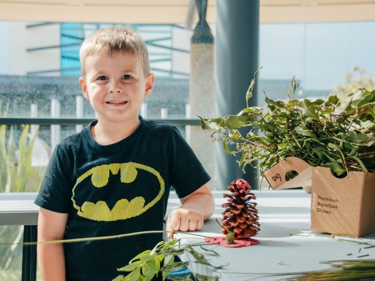 Image of A young boy wearing a Batman T-shirt smiles at a table outdoors. On the table are green leaves, a red-painted pine cone, and a cardboard box filled with branches.