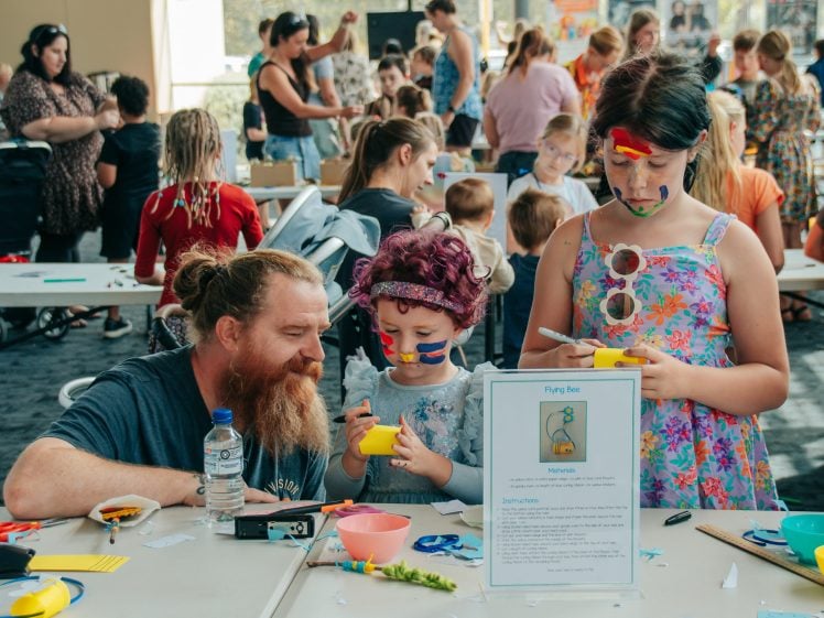 Image of A bearded man and two children with painted faces are crafting at a busy indoor event. The children hold yellow bowls, and a sign with instructions is visible on the table amidst art supplies and other participants.