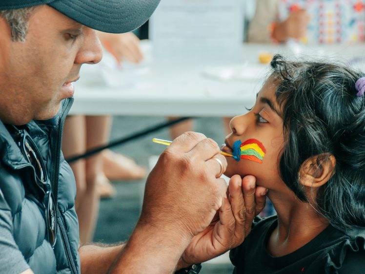 Image of A man carefully paints a rainbow on a young girl’s cheek at a table. The girl sits still, looking up at him, whilst other people are blurred in the background.
