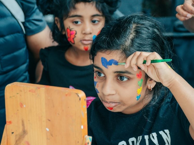 Image of Two young girls with colourful face paint look into a mirror. One girl holds a brush, painting a blue butterfly on her forehead, while her cheeks are decorated with flowers. The other girl has a red butterfly painted on her cheek.