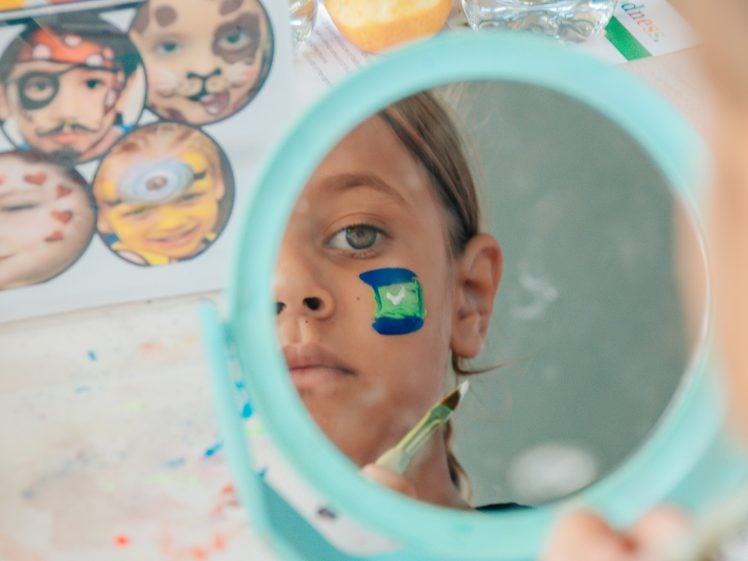 Image of A child looks in a handheld mirror while painting a colourful flag with a green centre on their cheek. Face paint supplies and a sheet of face paint designs are visible in the background.