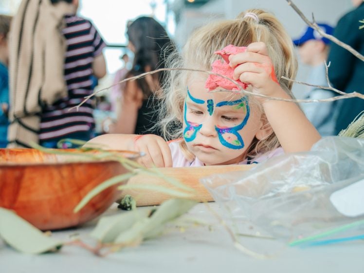 Image of A young child with a colourful butterfly painted on her face sits at a table, focussed on crafting with leaves and branches. Other children and adults are visible in the blurry background.