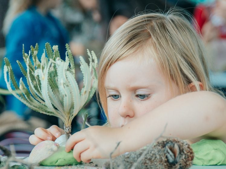 Image of A young child with blonde hair is focused on crafting, arranging natural materials like branches, rocks, and moss on a table. Other children and adults are visible blurred in the background.