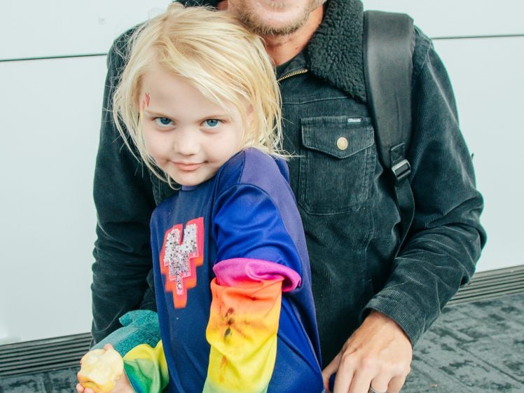 Image of An adult with colourful face paint smiles while kneeling beside a child in a blue rainbow outfit, also with face paint. They are indoors in front of white walls decorated with colourful artwork.