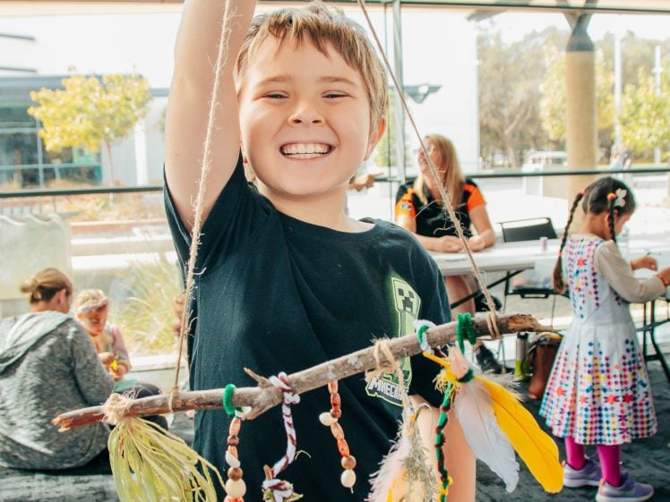 Image of A smiling child holds up a stick decorated with colourful beads, feathers, and string. Other children and adults are seated and working at tables in the background, in a bright indoor space with large windows.