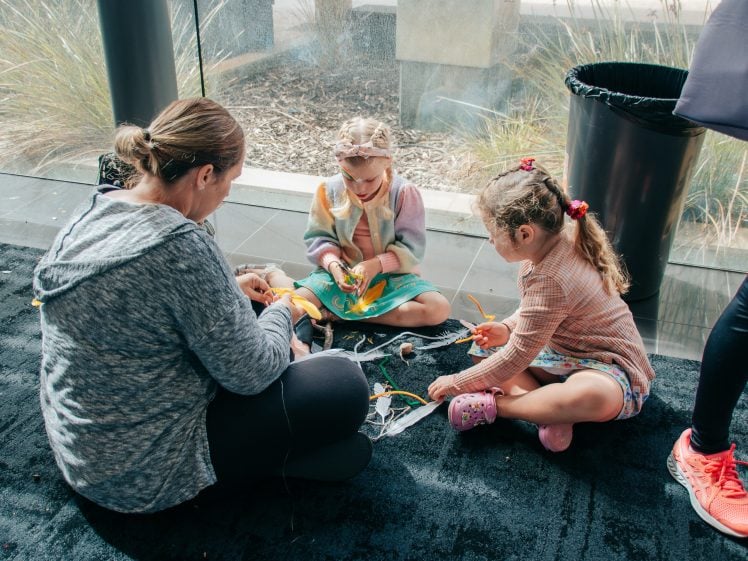 Image of A woman and two young girls sit on a carpeted floor indoors, making crafts together with colourful materials. Sunlight streams in from nearby windows, and a rubbish bin is visible in the background.