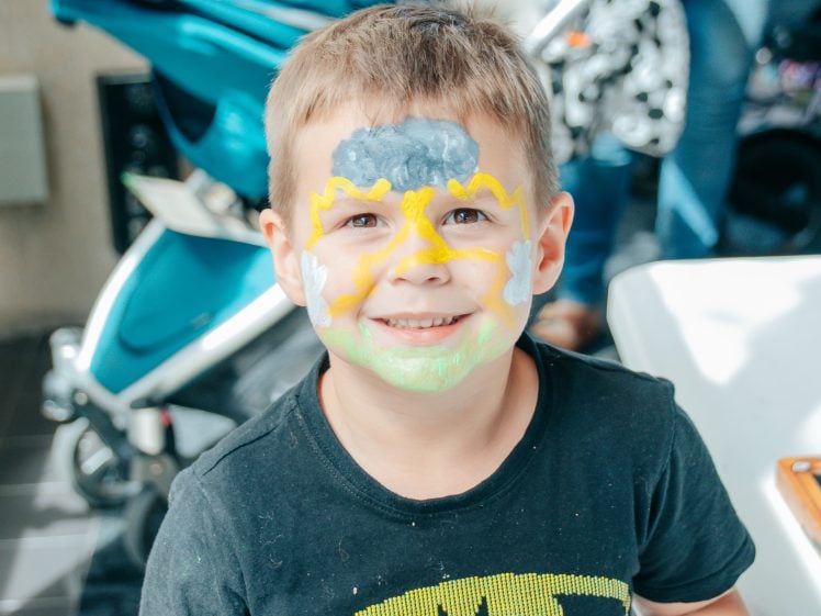 Image of A smiling young boy with face paint in yellow, green, white, and grey colours stands indoors. He wears a black T-shirt, and a blue pushchair is visible in the background.