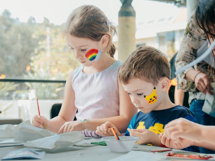 Image of Two children with painted faces, one with a rainbow and one with Pikachu, sit at a table focusing on arts and crafts activities. Adults and other children are nearby, and sunlight streams in from the background.