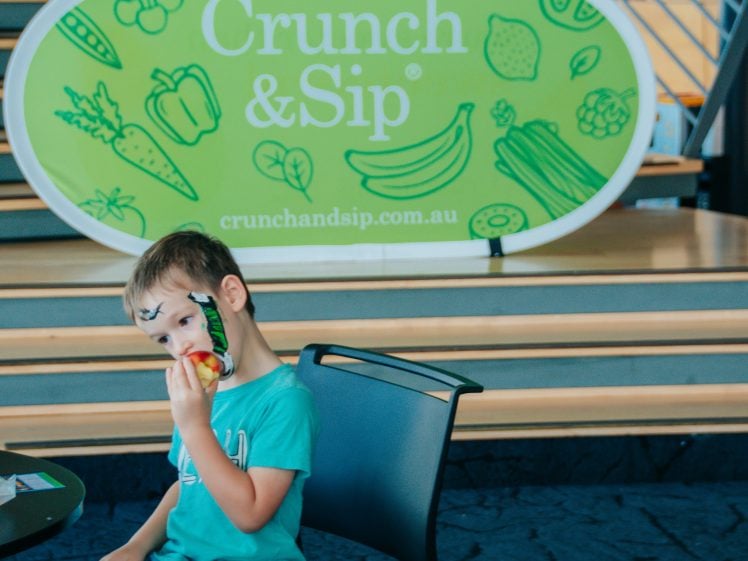 Image of A young boy with face paint eats an apple while sitting on a chair. In the background, there is a green Crunch & Sip sign with illustrations of fruit and vegetables.