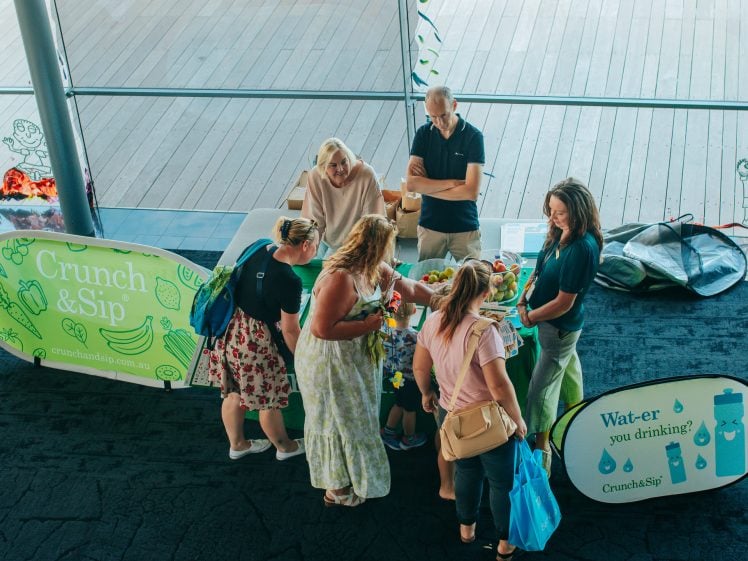 Image of A group of people gathers around a table with fruit and vegetables at a Crunch&Sip event. Two banners promote healthy eating and water, while staff assist attendees in a bright, modern indoor space.