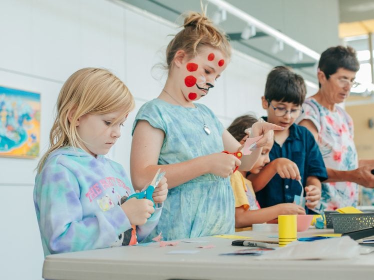 Image of A group of children and an adult stand at a table doing arts and crafts. One girl has red spots painted on her face. They are focused on cutting and arranging colourful paper in a bright, indoor setting.