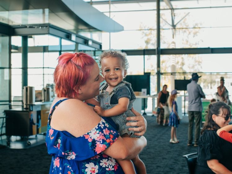 Image of A woman with pink hair in a blue floral dress holds a smiling toddler inside a bright, modern building. Other people, including children, are seen in the background.