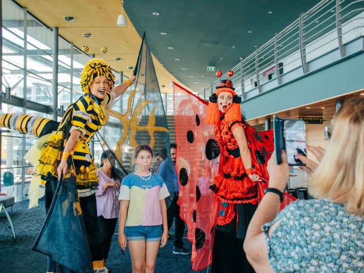 Image of Two performers on stilts dressed as a bee and a ladybird pose with children in a bright, modern indoor space while a woman takes their photo with her mobile.
