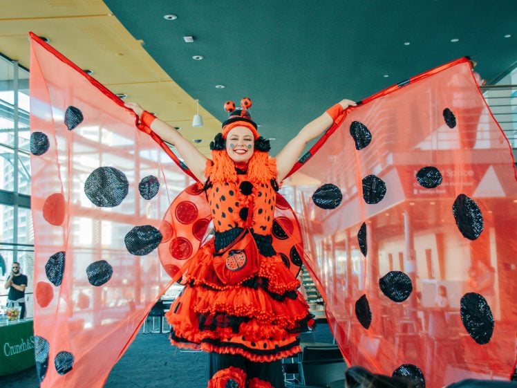 Image of A performer dressed in a bright, elaborate ladybird costume stands with arms raised, spreading large red wings with black spots inside a spacious, sunlit room.