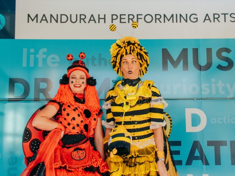 Image of Two adults in vibrant insect costumes pose in front of a Mandurah Performing Arts sign; one is dressed as a ladybird, the other as a bee, both smiling and making playful expressions.