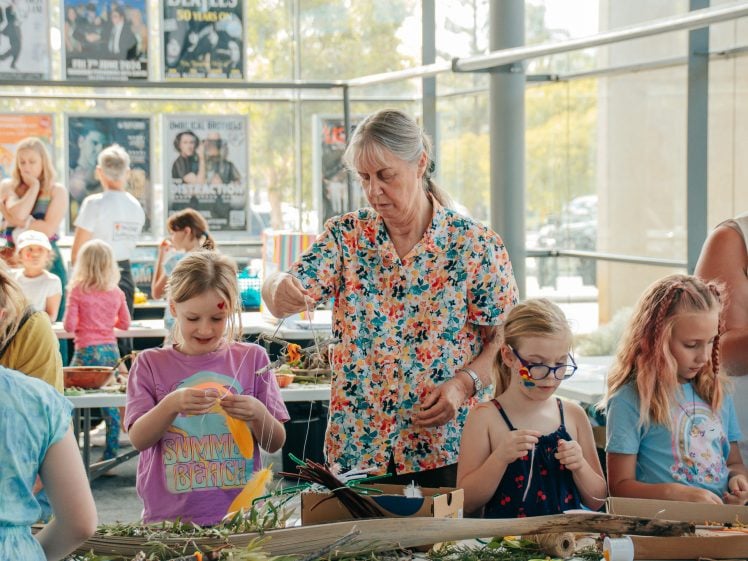 Image of A group of children and an older woman work together on arts and crafts at a table in a bright, glass-walled room. The older woman helps with materials whilst the children focus on their creative projects.