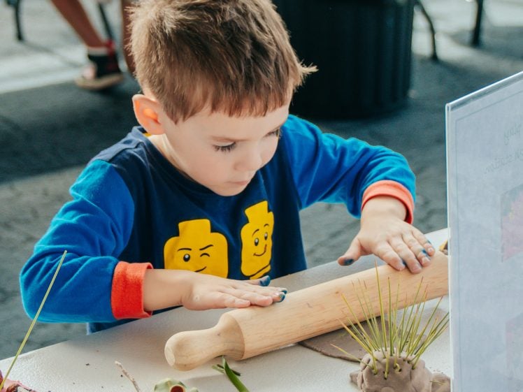 Image of A young boy in a blue shirt with Lego faces rolls dough with a rolling pin at a craft table, surrounded by natural materials like pine cones, sticks, and leaves. Other people and tables are visible in the background.