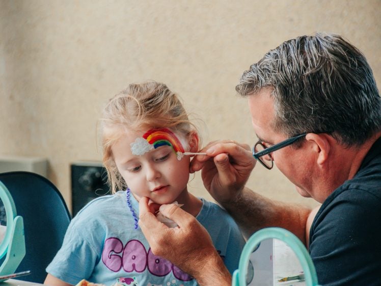 Image of A man carefully paints a rainbow with a cloud on a young girl’s forehead as she sits still, wearing a blue shirt and a beaded necklace, with mirrors and painting supplies around them.