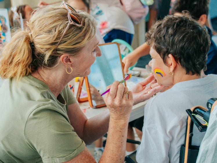 Image of A woman paints a rainbow on a smiling child's face at a table. The child is seated and looking in a mirror, whilst other children and colourful decorations are visible in the background.