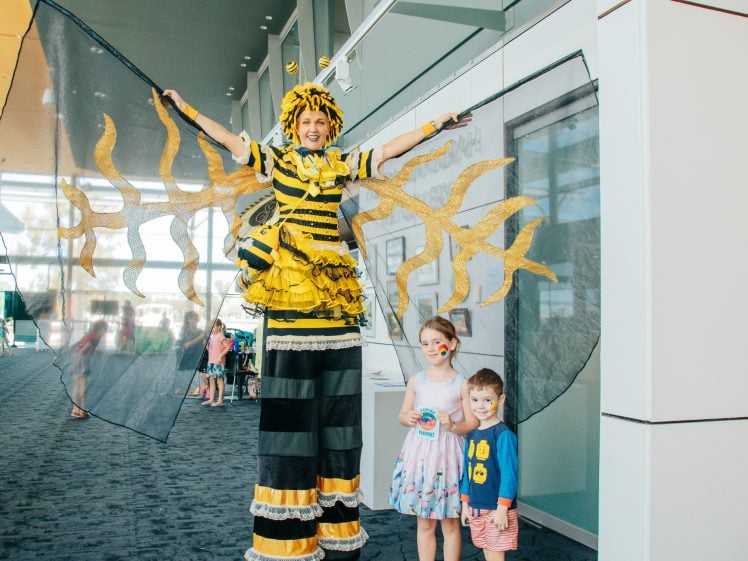 Image of A performer dressed as a bee with large wings stands on stilts indoors, smiling with arms outstretched. Two young children stand in front, looking at the camera; one has a butterfly painted on her face.
