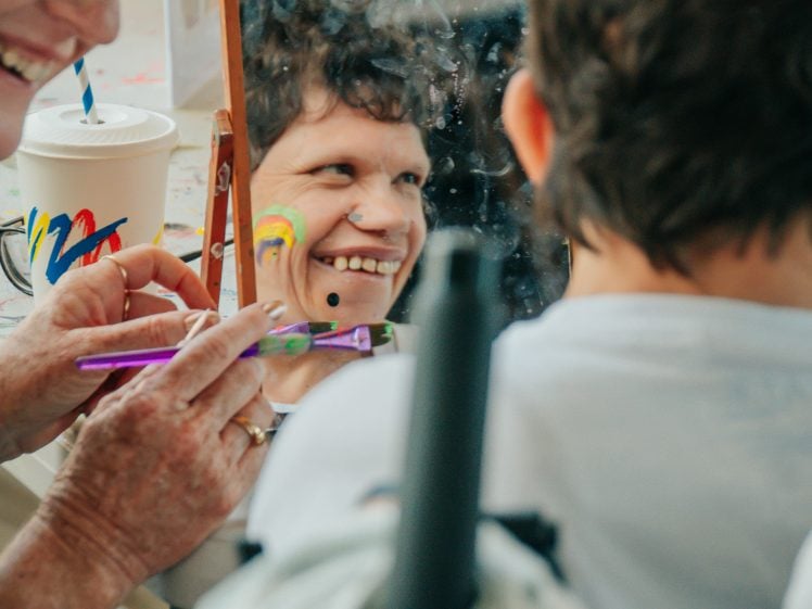 Image of A person smiles as their reflection in a mirror shows colourful face paint being applied by another person, with a cup and art supplies visible in the background.