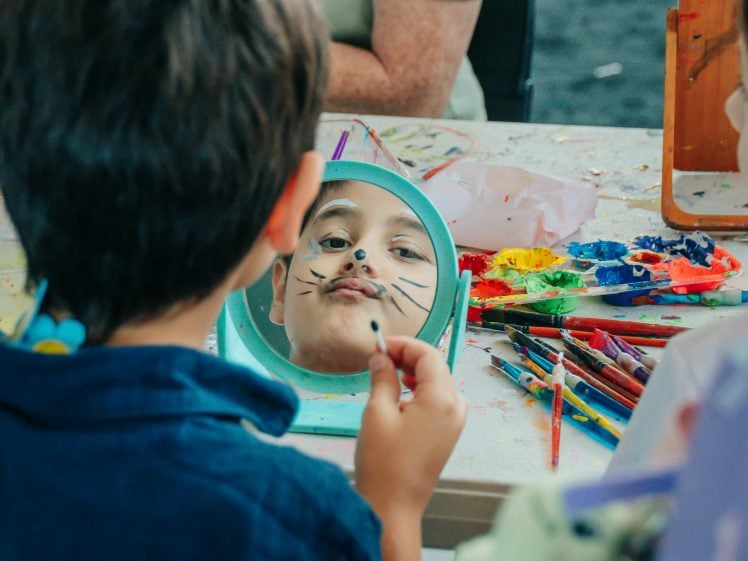 Image of A child looks in a mirror whilst another paints whiskers on their face. The table is covered with colourful paints, brushes, and art supplies, creating a playful and creative atmosphere.