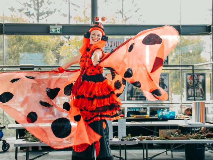 Image of A performer dressed in a bright red and black ladybird costume with large wings stands indoors, smiling and posing with arms outstretched. Tables and people can be seen in the background.
