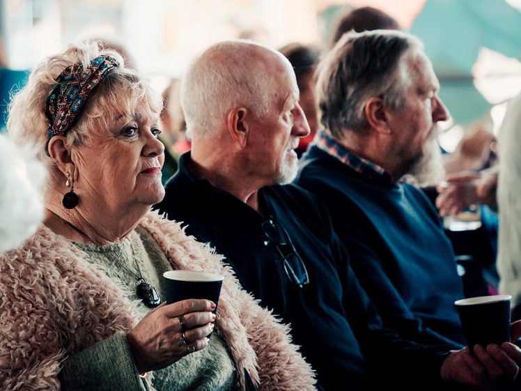 Image of Three older adults sitting side by side, holding cups and attentively watching something out of frame in a busy indoor setting. The woman at the front wears a headband and textured jacket.