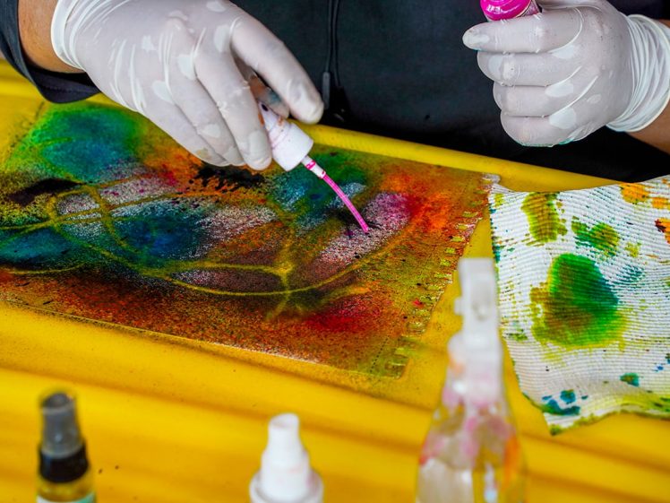 Image of A person wearing gloves uses a pink bottle to add colour to a vibrant, abstract painting on paper, with colourful stains on a cloth and spray bottles nearby.