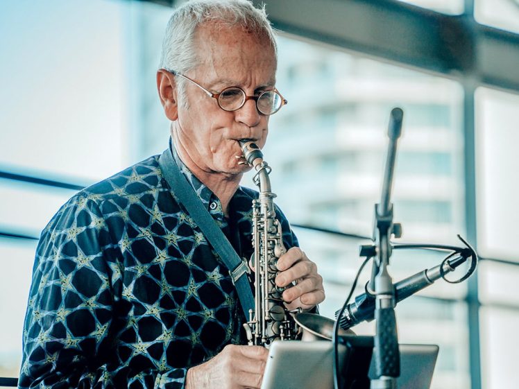 Image of An older man with glasses and grey hair plays a soprano saxophone indoors. He wears a patterned shirt and stands in front of microphones and a music stand, with large windows and a blurred building in the background.