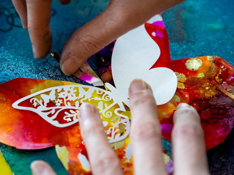 Image of Close-up of hands assembling a colourful paper butterfly with vibrant, mixed colours and patterns on a blue background; one hand holds the butterfly whilst the other arranges its intricate, white cut-out wings.