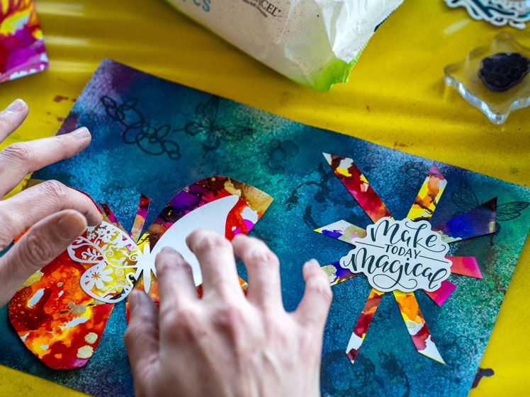 Image of A person arranges colourful cut-out shapes and the phrase “Make Today Magical” on a vibrant, textured blue and green art sheet whilst working on a creative craft project.