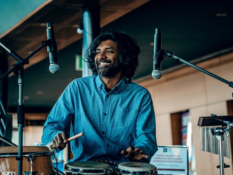 Image of A smiling person with long hair plays the bongos between two microphones on a stage, wearing a blue shirt. Other percussion instruments and a blurred indoor background are also visible.