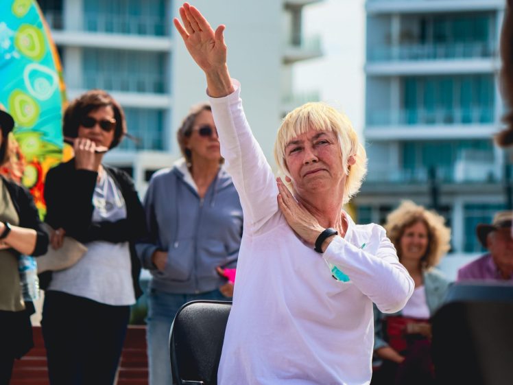 Image of An older woman in a white shirt sits outdoors, raising her right arm in the air whilst others watch in the background. The group appears engaged in an activity or performance, with buildings visible behind them.
