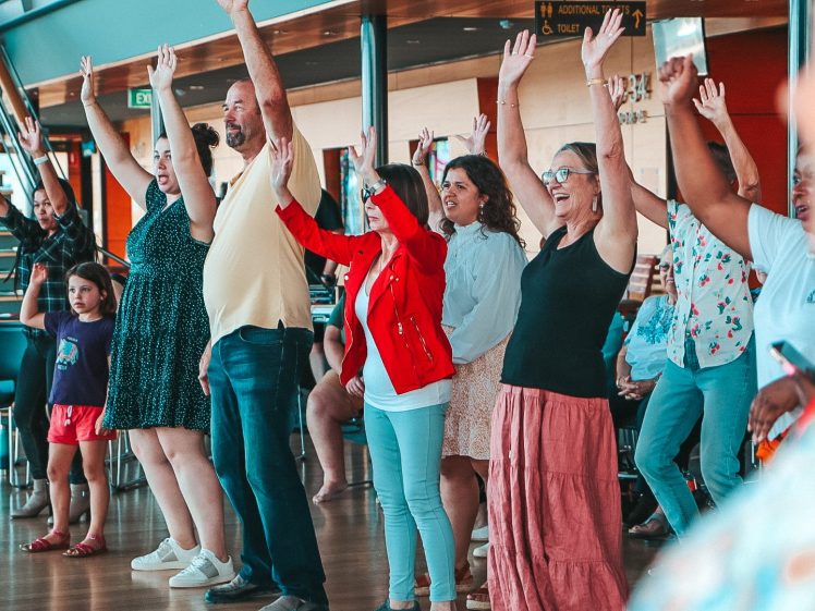 Image of A diverse group of people stand indoors with arms raised, smiling and participating in an activity. Some are standing, while others, including a child in purple, are nearby. The setting appears to be a community or recreational space.