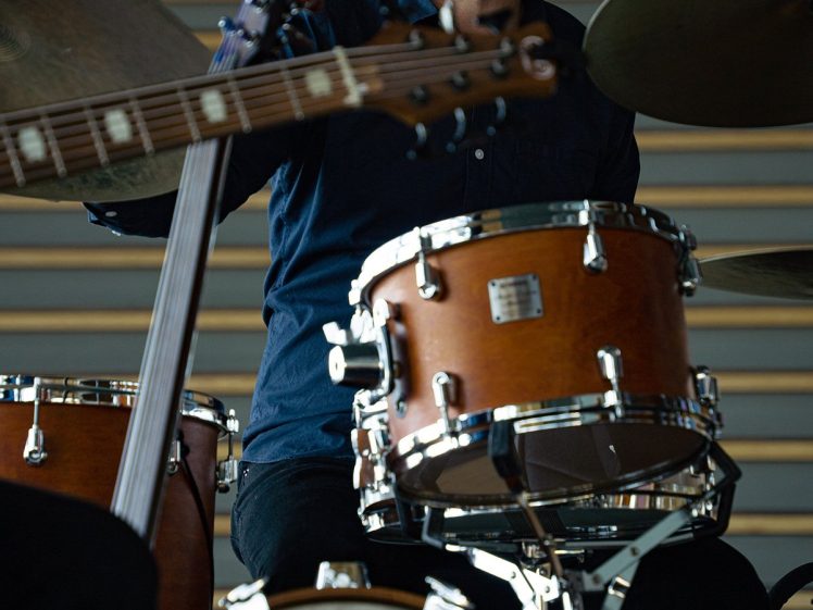 Image of A bearded man in a dark shirt plays a drum kit on stage, with a guitar partially visible in the foreground and tiered seating in the background.